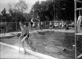 A boy is emerging from a pool in the foreground while several boys in the background are standing around the pool and two others are in the midst of diving and jumping into the pool.